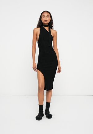 Black ribbed knit dress with a halter neckline, open back, and a high slit on the left side; paired with black sandals and socks.