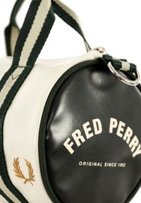 Bolso blanco y negro con acentos verdes, que presenta materiales texturizados, asas y una base circular. Logotipo "FRED PERRY" y emblema visibles.