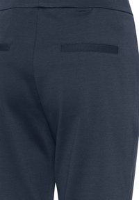 Sötét tengerészkék nadrág, texturált felülettel, két vízszintes hátsó zsebbel, melyek varrott díszítéssel vannak rögzítve.