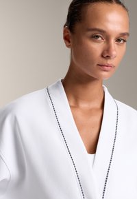 Bela obleka s šalom, ki ima temno obrobo po robovih. Gladka tekstura in minimalistična zasnova.