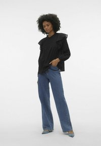 Blusa nera con spalle arricciate, tessuto testurizzato e vita arricciata, abbinata a jeans a gamba larga blu e décolleté punta leggera blu.