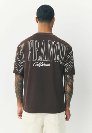 Braunes Baumwoll-T-Shirt mit fettem weißen Text "SAN FRANCISCO" und "California" auf dem Rücken, lockere Passform, kurze Ärmel und dezente Textur.