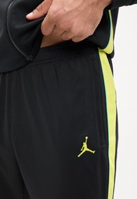 Mano ajustando la cremallera de una chaqueta negra, vistiendo pantalones negros con una franja lateral amarillo-verde y un logo Jumpman amarillo neón cerca del bolsillo.