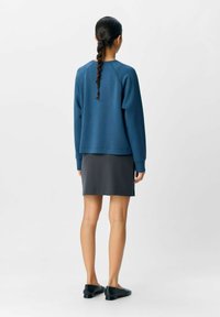 Blauer Langarm-Sweater mit lockerer Passform und strukturiertem Stoff, kombiniert mit einem grauen Rock. Die Schuhe sind schwarze Ballerinas mit niedriger Profil.