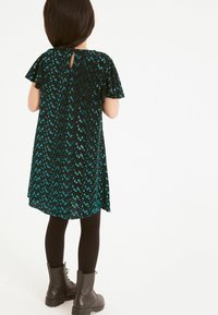 Robe noire à manches courtes avec des motifs en sequins turquoise en chevrons ; possède un dos en forme de clé et est associée à des leggings noirs et des bottes.