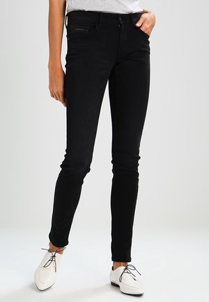 Vrouw draagt strakzittende zwarte spijkerbroek met vijf zakken en witte veterschoenen, staand tegen een effen achtergrond.