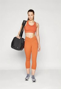 Γυναίκα με πορτοκαλί αθλητικό σουτιέν και κολάν, κρατώντας μαύρη αθλητική τσάντα στον ώμο, φορώντας γκρί αθλητικά παπούτσια, στέκεται μπροστά σε λευκό φόντο.