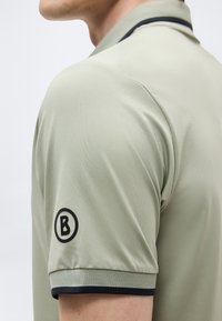 Gros plan sur une personne portant un polo gris clair avec un liseré noir sur le col et la manche, arborant un patch logo "B" noir sur la manche.