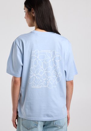 Vrouw met lang donker haar die een lichtblauw T-shirt draagt met een wit abstract bloemenpatroon en tekst op de achterkant, gecombineerd met lichte spijkerbroeken.
