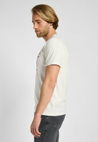 Camiseta de color beige claro hecha de tela suave, con mangas cortas y escote redondo. El gráfico frontal incluye texto y diseños simples.