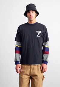 Black short-sleeve t-shirt with white "OBEY has a Posse" text, layered over a multicolored long-sleeve shirt, worn with khaki pants.