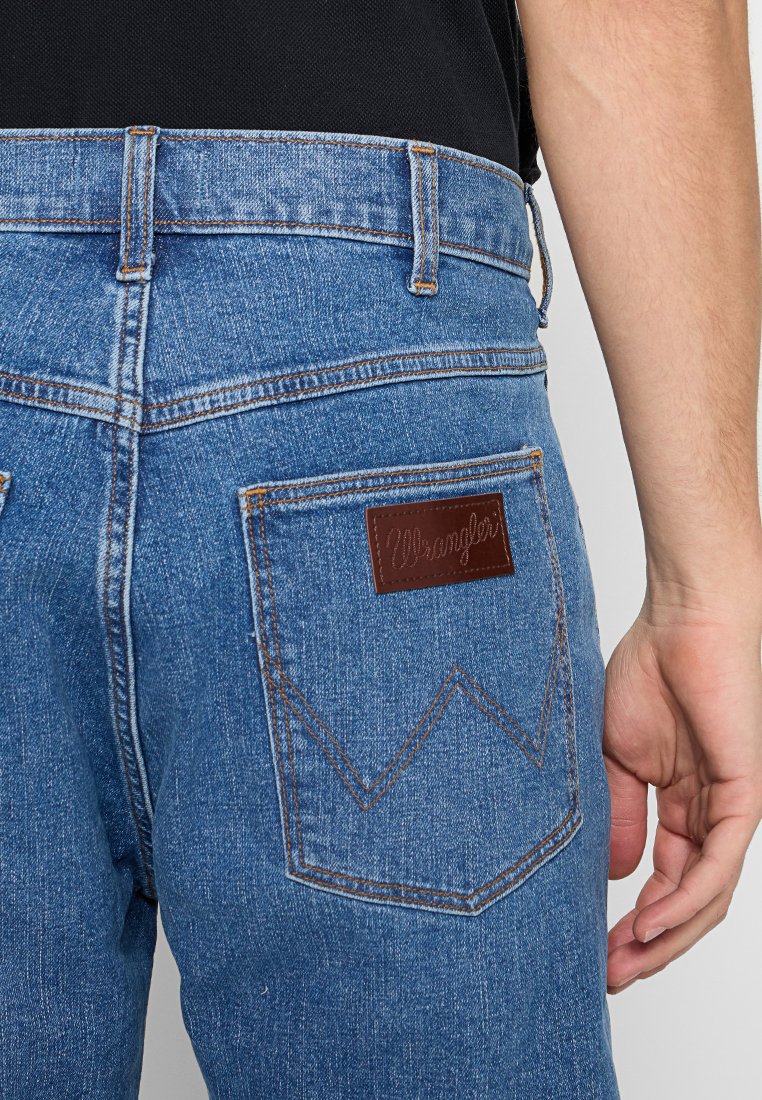 Niebieskie dżinsy z wysokim stanem, z brązową skórzaną łatą z logo "Wrangler" oraz kontrastowymi pomarańczowymi szwami na kieszeniach.