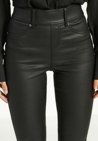 Pantaloni attillati neri in similpelle con tasche frontali e passanti per cintura, indossati con un top nero a maniche lunghe, le mani poggiate vicino alle cosce.
