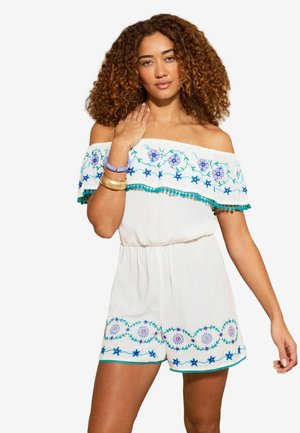 Žena s kučeravými vlasmi v bielom overal s odhalenými ramenami, zdobenom modrým a purpurovým kvetinovým výšivkom, a modrými náramkami na ľavom zápästí.