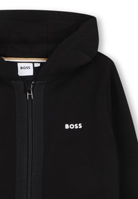Sweat à capuche noir zippé avec capuche et logo "BOSS" blanc sur le côté gauche de la poitrine, présenté de face sur la partie supérieure.