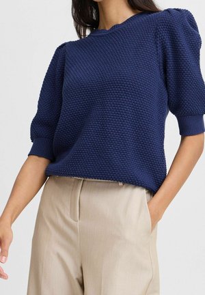 Kvinde iført marineblå tekstureret kort puffærmet sweater og beige højtaljede bukser med hånden i lommen mod ensfarvet baggrund.