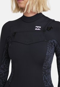 Svart våtdräkt med hög krage, utrustad med en ficka med dragkedja och texturerade sidopaneler med abstrakta mönster. Logotypen visas på bröstet.