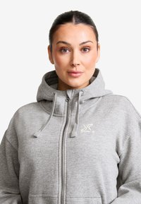 Grå zip-up huvtröja med huva, tillverkad av mjukt material. Har en frontdragkedja, snoddar och logotypdetalj på bröstet.
