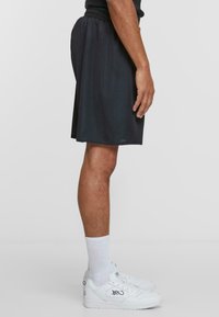 Sorte treningsshorts i mesh med elastisk liv, løs passform og sidelommer. Brukt med hvite joggesko og hvite ankelsokker.