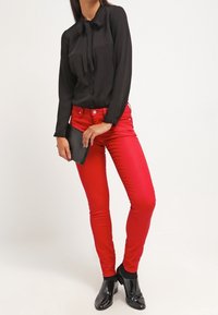 Czarna bluzka z długim rękawem i wiązaniem przy szyi, w zestawieniu z dopasowanymi czerwonymi dżinsami i czarnymi lakierowanymi botkami za kostkę. Trzyma czarną kopertówkę.
