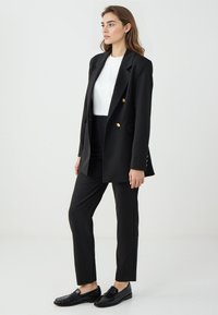 Zwarte blazer met gouden knopen, witte top, op maat gemaakte zwarte broek en zwarte instappers. Soepele textuur, gestructureerd ontwerp, moderne pasvorm.