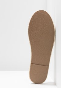 Semelle de chaussure espadrille beige avec fond en caoutchouc texturé et détail de coutures le long du bord, présentée verticalement sur un fond blanc.