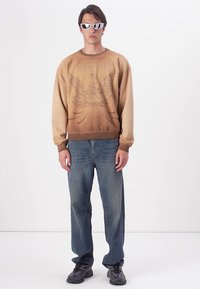 Bruine sweatshirt met een grafisch ontwerp van een adelaar, gecombineerd met lichtblauwe jeans en zwarte schoenen. Model draagt witte zonnebril.