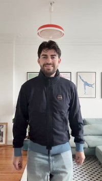 Tamsiai mėlyna zip-up striukė su aukštu apykakle, raištelių rankovėmis ir kontrastinga šviesiai mėlyna juostos dalimi. Ant krūtinės yra apvalus logotipas.