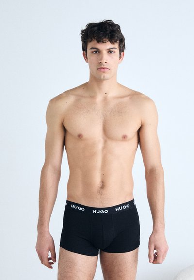 Caleçons boxers noirs avec un logo blanc "HUGO" sur la ceinture, fabriqués en tissu lisse, design ajusté, vue de face et de côté d'un modèle masculin.