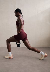 Rød mønstrete sports-bh og shorts, holder en svart kettlebell; iført nøytrale treningssko med fargede detaljer og hvite sokker.