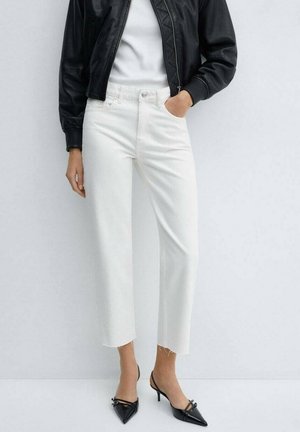 Mujer con vaqueros blancos recortados con dobladillos sin rematar, tacones negros de punta afilada con tira trasera, top blanco y chaqueta de cuero negra, una mano en el bolsillo.
