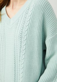 Femme portant un pull en maille torsadée bleu clair avec col en V et motifs texturés, visible des épaules à la taille.