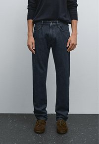 Mörkblå denimjeans med rak bendesign, fem fickor och knappstängning. Bärs med bruna mockaskor.