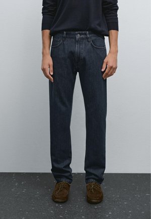 Dark blue denim jeans with a straight leg design, five pockets, and a button closure. Worn with brown suede shoes.