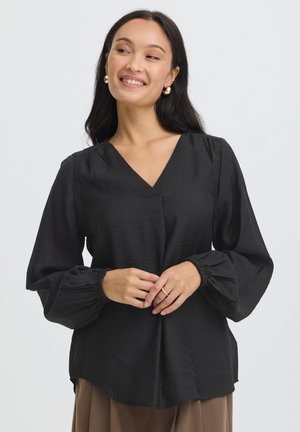 Kvinde med langt mørkt hår iført gyldne øreringe, en sort bluse med lange ærmer med pufærmer og brune bukser, smilende og ser til siden.