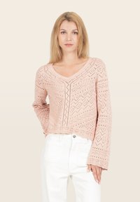 Pull en tricot rose avec un col en V, des manches longues et des motifs ajourés complexes. Fini avec un ourlet texturé. Porté sur un pantalon blanc.