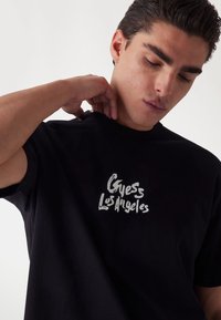 Camiseta de algodón negra con un logo gráfico blanco "Guess Los Ángeles" en el pecho. Diseño de cuello redondo y corte relajado.