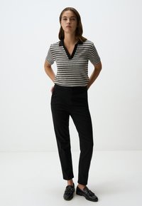 Polo shirt a righe in maglia con colletto a V nero, abbinato a pantaloni neri a sigaretta e mocassini neri, su uno sfondo bianco semplice.