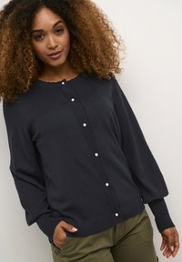 Tumši zili jaciņas ar apaļu kakla izgriezumu, teksturētu diegu šuvēm un pērļu pogām pa priekšu. Iekļauj garas, pufīgas piedurknes.