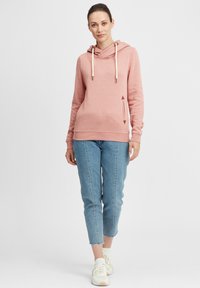 Felpa rosa con cappuccio e collo a tazza, con lacci e tasca laterale. Abbinata a jeans azzurri a vita alta e scarpe da ginnastica bianche.