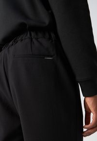 Personne portant un pantalon noir avec une taille élastique et une petite étiquette métallique de marque au-dessus de la poche arrière, à côté de sa main droite détendue.