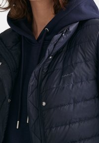 Marinblå vadderad väst med diamantquiltat mönster, med dragkedja fram, tryckknappar och en subtil logotyp på tyget. Bärs över en marinblå hoodie.