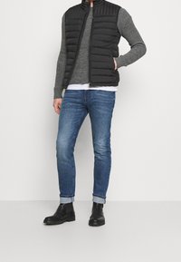 Svart puffig väst över en grå stickad tröja, kombinerad med blå tajta jeans och svarta ankelstövlar. Vesten har horisontell sömnad.