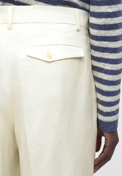 Κοντινό πλάνο της πίσω τσέπης με κουμπί σε λευκό παντελόνι, που φοριέται με μπλε και άσπρο ριγέ πλεκτό μακρυμάνικο πουκάμισο.