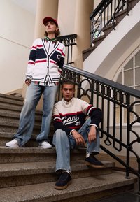 Two individuals on a staircase. One wears a striped cardigan and cap; the other is in a crewneck sweater. Both in loose denim and sneakers.
