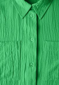 Camicia verde con texture, colletto button-down, bottoni visibili e due tasche frontali con una lavorazione del tessuto stropicciata.