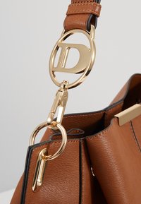 Sac à main en cuir marron avec fermeture métallique dorée et logo circulaire doré comportant la lettre D attaché à la lanière de la poignée.