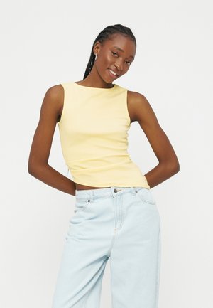 Jeune femme souriante, portant un haut sans manches jaune pâle et un jean taille haute bleu clair, debout avec les mains derrière le dos.