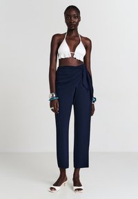 Femme portant un haut crop blanc à bretelles, un pantalon bleu marine style enveloppant, des sandales à talons blancs et des bracelets argentés et bleus aux deux poignets.