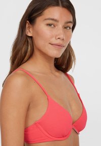 Coral pink padded bra with thin adjustable straps. Textured fabric with a smooth finish and a subtle logo detail on the band.